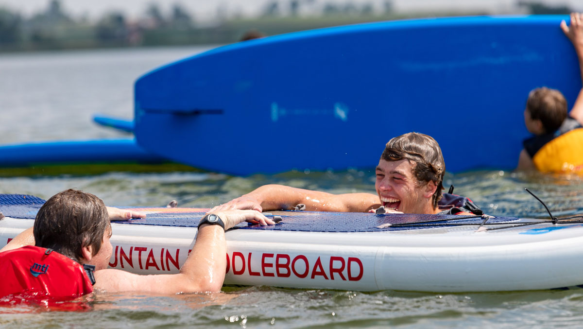 Two teenage campers smile in the water while trying to get on a stand up paddle board