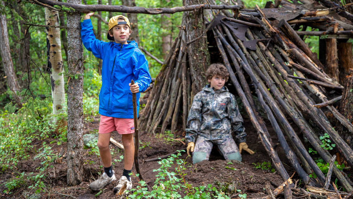 Two young campers stand in the forest by a shelter made from branches